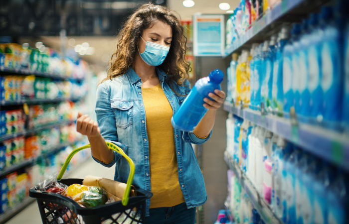 Young female shopping in grocery store, holding a basket in one hand and dish detergent in another hand