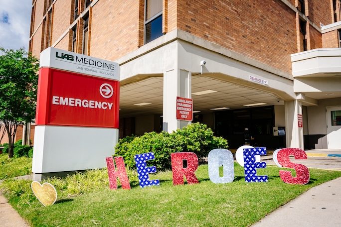 Exterior of UAB Hospital-Highlands showing the Emergency entrance with decorative lettering that reads "Heroes" staked in the grass in patriotic colors during the COVID-19 (Coronavirus Disease) pandemic; blue sky and white clouds are above, April 2020.